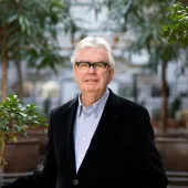 A grey-haired Sir Terry Farrell (1938-2025) wearing glasses, a dark jacket and light blue shirt photographed in front of an avenue of small trees in an urban setting © richard gleed / farrell