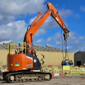 An orange, zero tail-swing midi excavator lifts a pile cropper on a construction site
