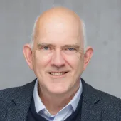 Head and shoulders photo of a bald, smiling man in open necked shirt, jumper and jacket