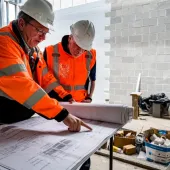 Two construction workers look at plans laid out on a table inside an under construction building