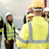 A group of youg people wearing yellow PPE and white and yellow hard hats listen to an older worker