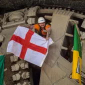A man wearing a hard hat and waving a Cross of St George flag pokes his head through a TBM cutter wheel with a Welsh flag and another unidentified flag adorning the cutter head as HS2 engineers celebrate the breakthrough of tunnel boring machine Elizabeth at Washwood Heath in Birmingham, completing the 3.5-mile Bromford tunnel under the M6. (Image © HS2 Ltd)