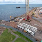 A red and yellow crawler crane lifts precast concrete elements at a shoreline construction site