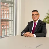 Vijay Bange in a dark suit, white shirt and red tie sits at an empty table with windows behind him showing a red brick building.