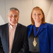 A smiling man and woman in business attire (dark suit, open necked shirt, blue jacket, black top) look at the camera