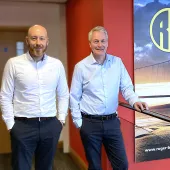 Two middle-aged men in smart, business casual wear stand on a stair landing in front of a large photo