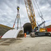 A yellow crawler crane lifts a precast concrete tunnel segment ready for placing on a cut and cover tunnel in the background