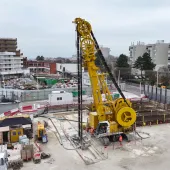An elevated view of a yellow Soilmec SC-110 Urban cutter working on the Grand Paris Express, constructing 1,200mm-thick diaphragm walls up to 38 metres deep at Rueil–Suresnes Mont-Valérien station, surrounded by houses, low rise buildings and roads.