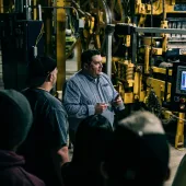 A bearded man, surrounded by a small group of people all looking at an LCD screen in a factory surrounded by heavy equipment