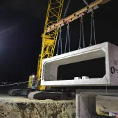 A yellow Kobelco crawler crane lifts a precast, rectangular concrete element