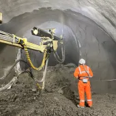 A construction worker in orange PPE uses a remote control unit to control a Normet concrete sprayer in a tunnel
