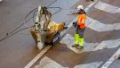 A man in orange, grey and yellow PPE uses a remote controlled piece of construction equipment fitted with a breaker to carry out repairs to a concrete road
