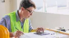 A man in glasses, blue shirt and yellow PPE vest sits working at a desk.