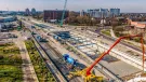 An aewrial view of a large construction site with a yellow and red concrete pump and blue mixer truck in the foreground, a blue crawler crane in the distance and against a city scape