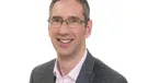 A head and shoulders shot of Peter Reekie of the Scottish Futures Trust will speak on sustainable infrastructure at the Glasgow Industry Day wearing glasses, an open necked pick shirt and a grey over-check jacket