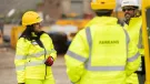 Three people in yellow PPE stand talking with construction machinery in the background