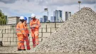 Three people in orange PPE stand discussing aggregate needs in a construction materials yard with a wall and city scape in the background