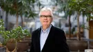 A grey-haired Sir Terry Farrell (1938-2025) wearing glasses, a dark jacket and light blue shirt photographed in front of an avenue of small trees in an urban setting © richard gleed / farrell