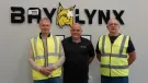 Three men stand in front of the BayLynx logo wearing PPE