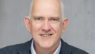 Head and shoulders photo of a bald, smiling man in open necked shirt, jumper and jacket
