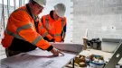 Two construction workers look at plans laid out on a table inside an under construction building