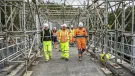 A group of construction workers walk along a concrete walkway, through a "tunnel" of scaffolding, wearing a variety of PPE , including orange hi vis jackets, yellow trousers, white hard hats and eye protectors