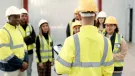 A group of youg people wearing yellow PPE and white and yellow hard hats listen to an older worker