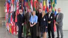 Four men and two women wearing work clothes stand in front of a large group of flags