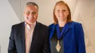 A smiling man and woman in business attire (dark suit, open necked shirt, blue jacket, black top) look at the camera