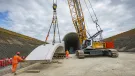 A yellow crawler crane lifts a precast concrete tunnel segment ready for placing on a cut and cover tunnel in the background