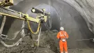 A construction worker in orange PPE uses a remote control unit to control a Normet concrete sprayer in a tunnel